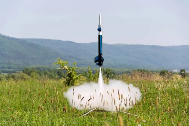 A blue and white colored model rocket launches in a cloud of smoke in the middle of field surrounded by rolling hills.