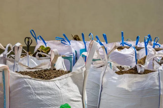 A group of white bags with blue stitching and handles gathered together and filled to the brim with potting soil.