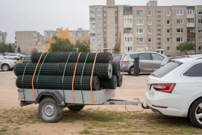 A parked white car with a trailer hitch and a utility trailer behind it carrying rolls of metal fencing strapped to the trailer.