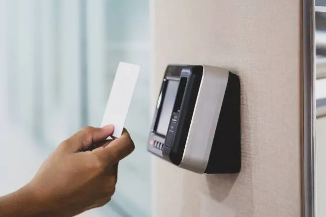 Someone standing outside a building holding a proximity key card up to the digital reader. The key card is white.