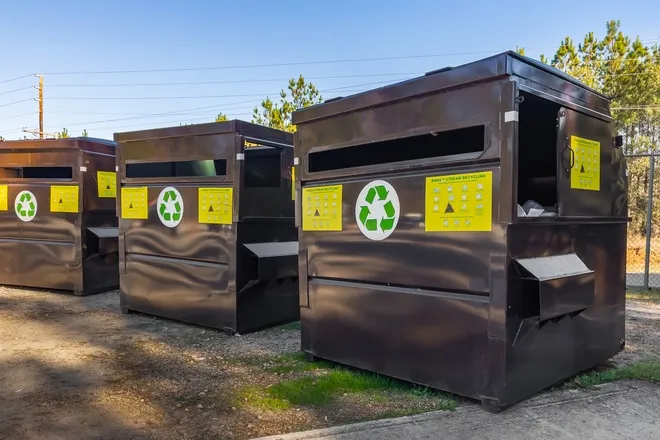 Large commercial recycling hoppers sitting outside. There are three gray hoppers with green recycling logos.