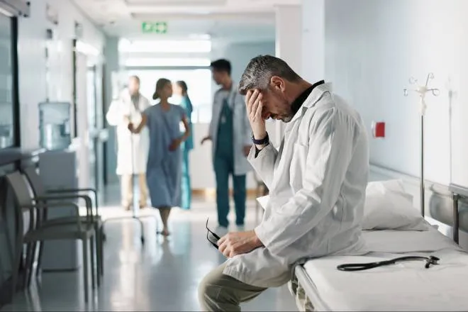 A doctor holding his head in one hand and his glasses in the other, resting on a gurney as other doctors and patients talk in the background.