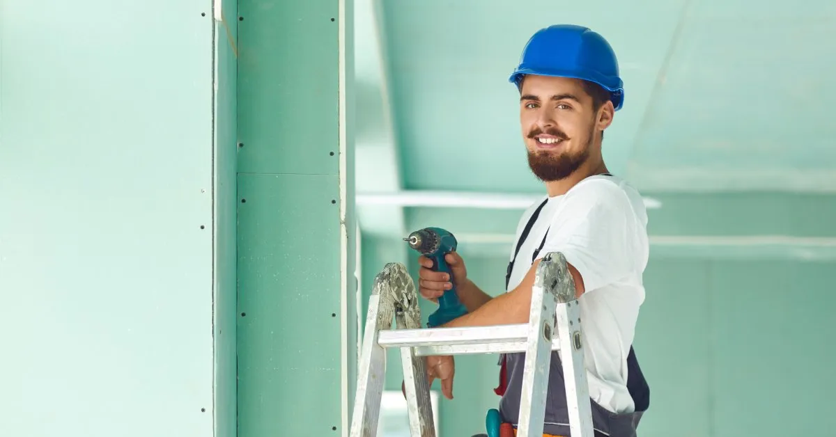 A male worker with a beard and mustache stands at the top of a ladder using a drill to install drywall sheets.