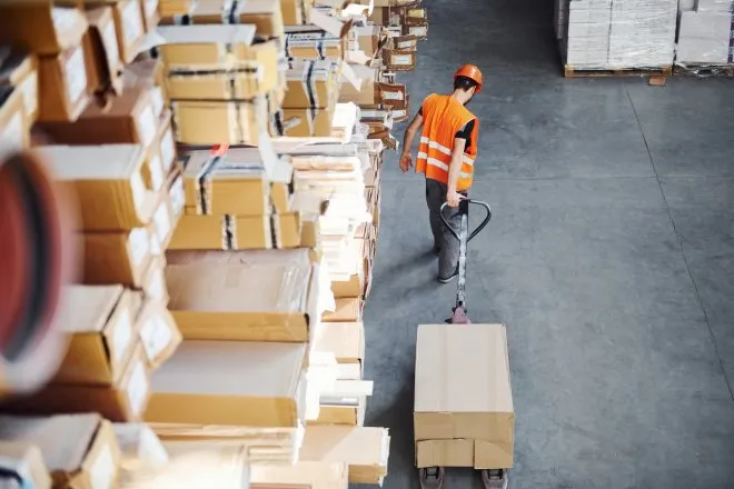 Aerial view of a warehouse worker pulling a cardboard box on a pallet truck next to shelving full of similar boxes and packages.