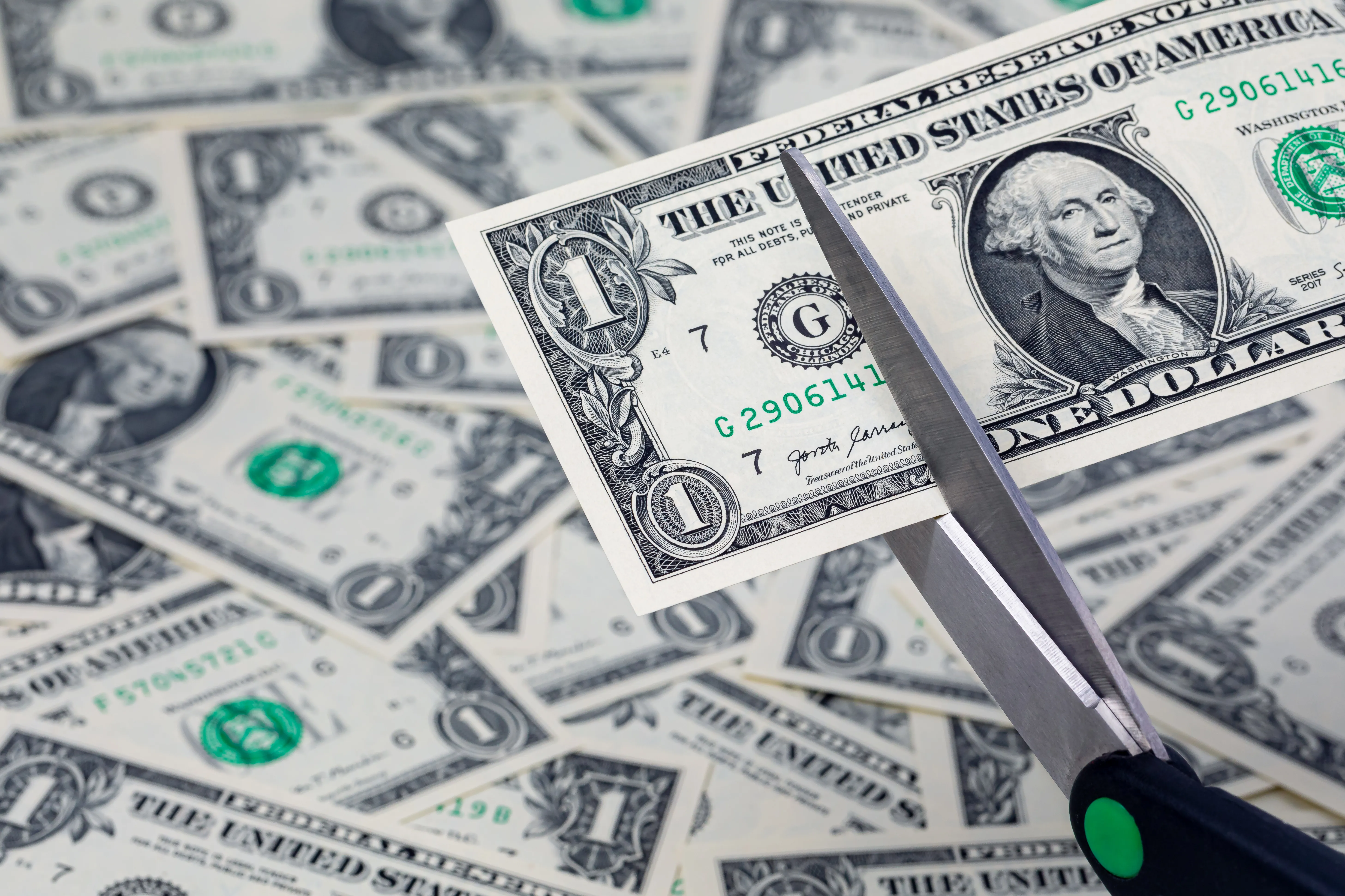 A pair of scissors is preparing to cut a United States one dollar note. In the background, additional dollar bills are strewn haphazardly.