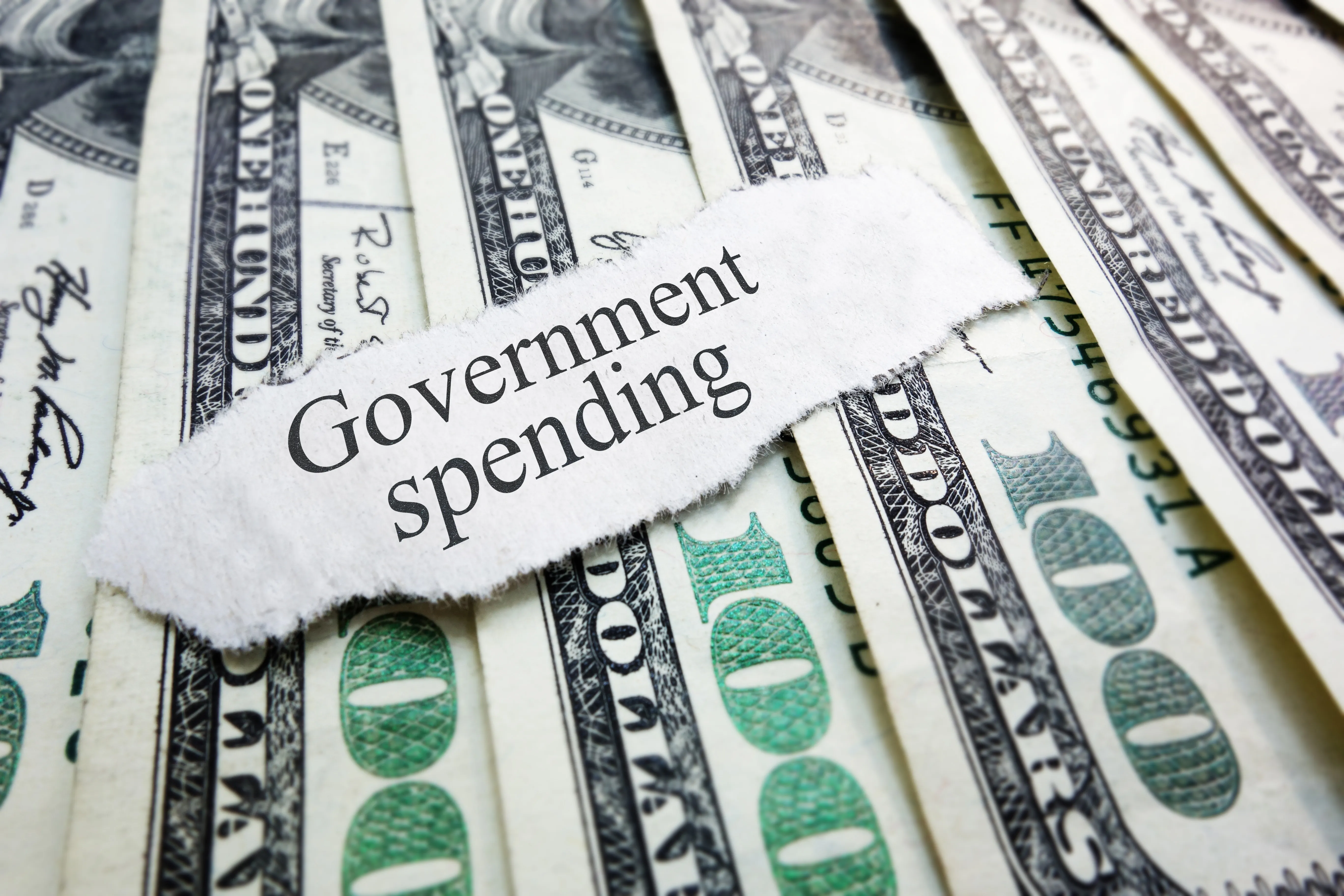 A scrap of torn paper with the words "Government spending" rests on top of a spread of United States 100 dollar currency.