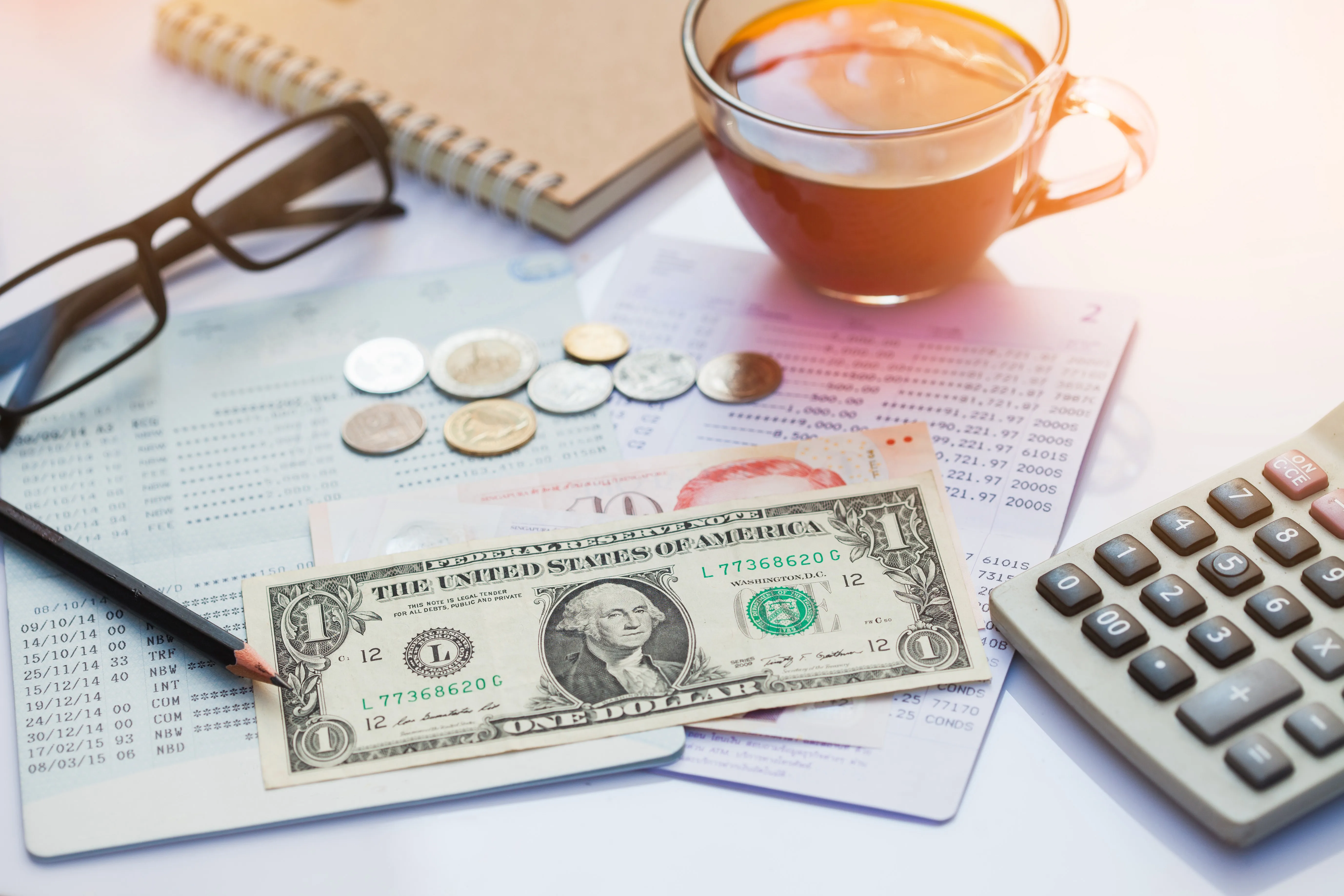 Financial statement with assorted United States currency and coins, calculator, eyeglasses, and a cup of coffee on top.