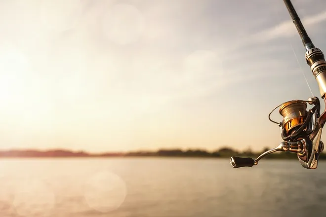 A close-up view shows a fishing pole with a brown handle. A body of water surrounded by trees in the background.
