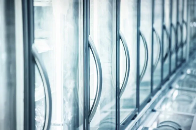 A close-up view shows a line of built-in refrigerators at a supermarket. The doors are glass, and the handles are silver.