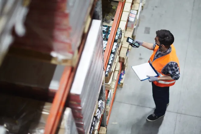A man in an orange vest is holding an electronic device up to a shelf filled with pallets with various items on them.
