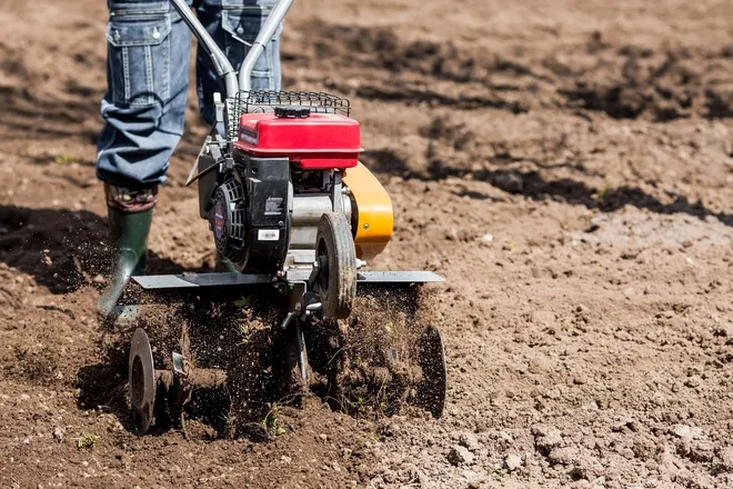 A person in farm attire walking a plot of land with a walking tiller. They're tilling the soil and wearing green boots.