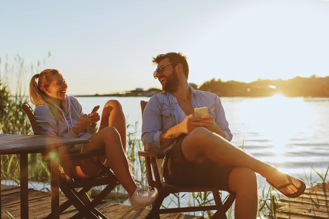 A young couple smiles and laughs as they sit on the side of a river. Both of them hold smartphones in their hands.