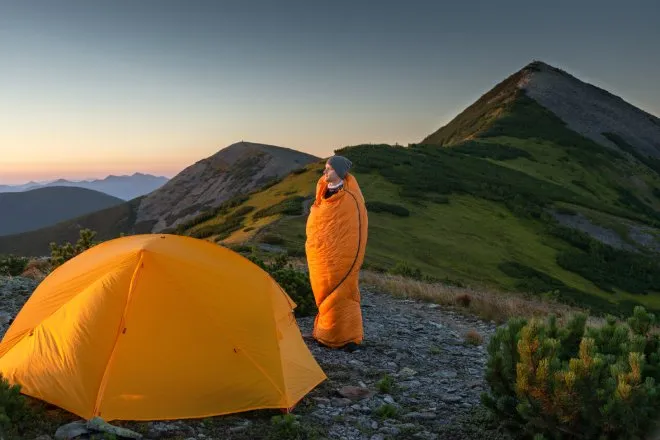 A man wraps himself in his orange sleeping bag as he gazes at the mountainous landscape beside his tent.