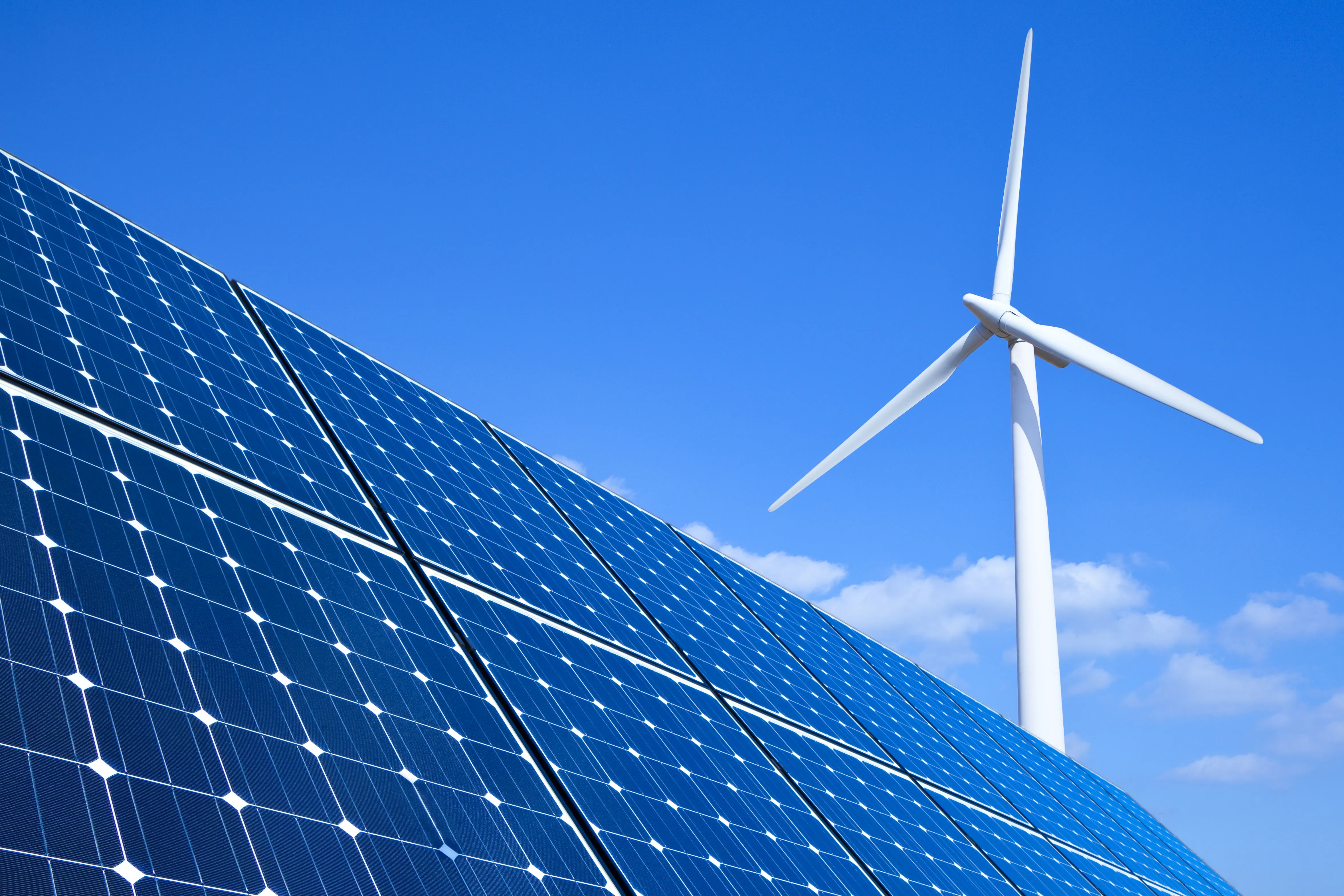 Solar panels in the foreground, with a wind turbine in the background, against a blue sky with a few clouds.