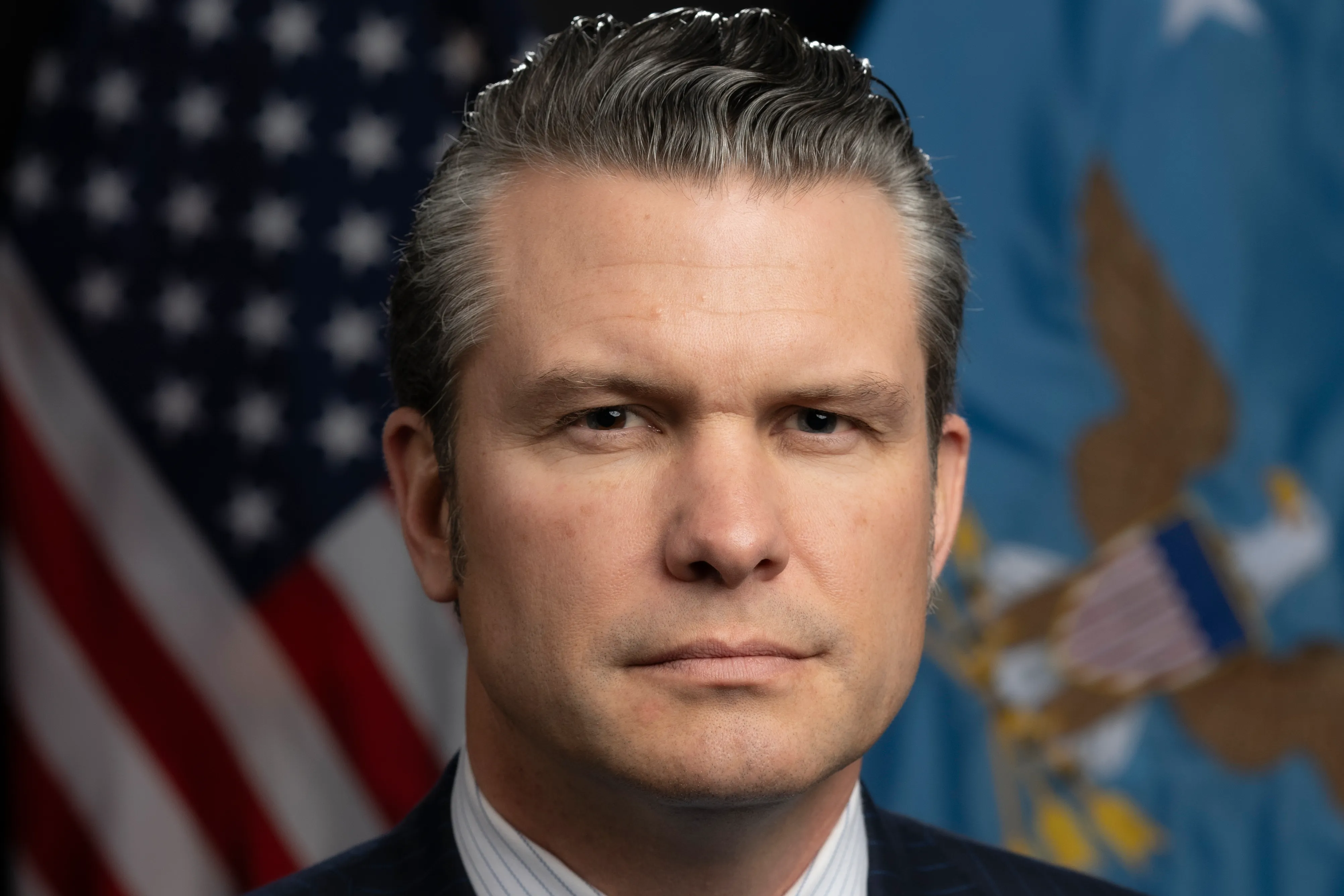 Head shot of Pete Hegseth, Secretary of Defense under the second Trump administration, in front of the flag of the United States and a military flag.