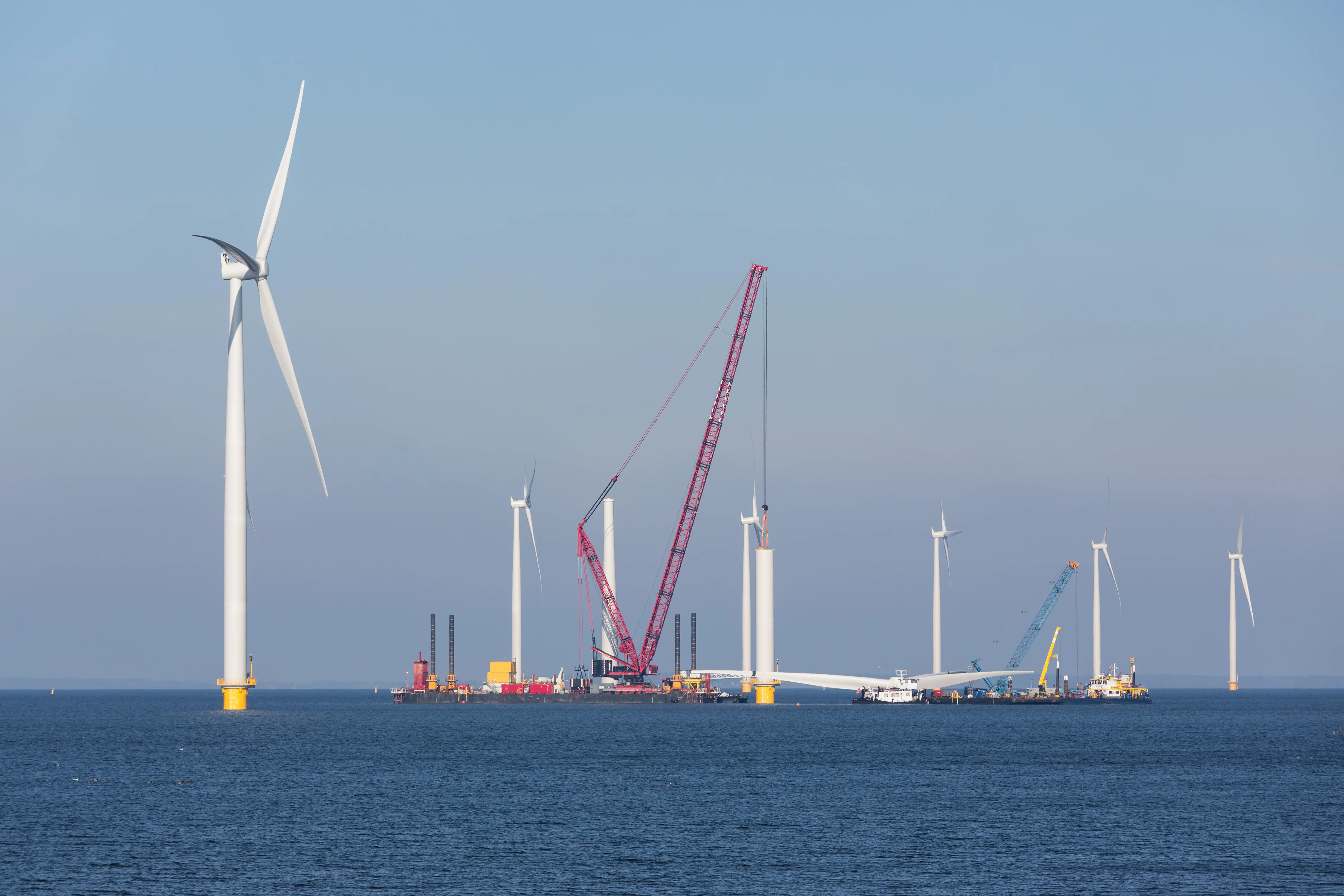 Wind turbines under construction in the ocean. A crane ship is in place to lift a blade assembly.