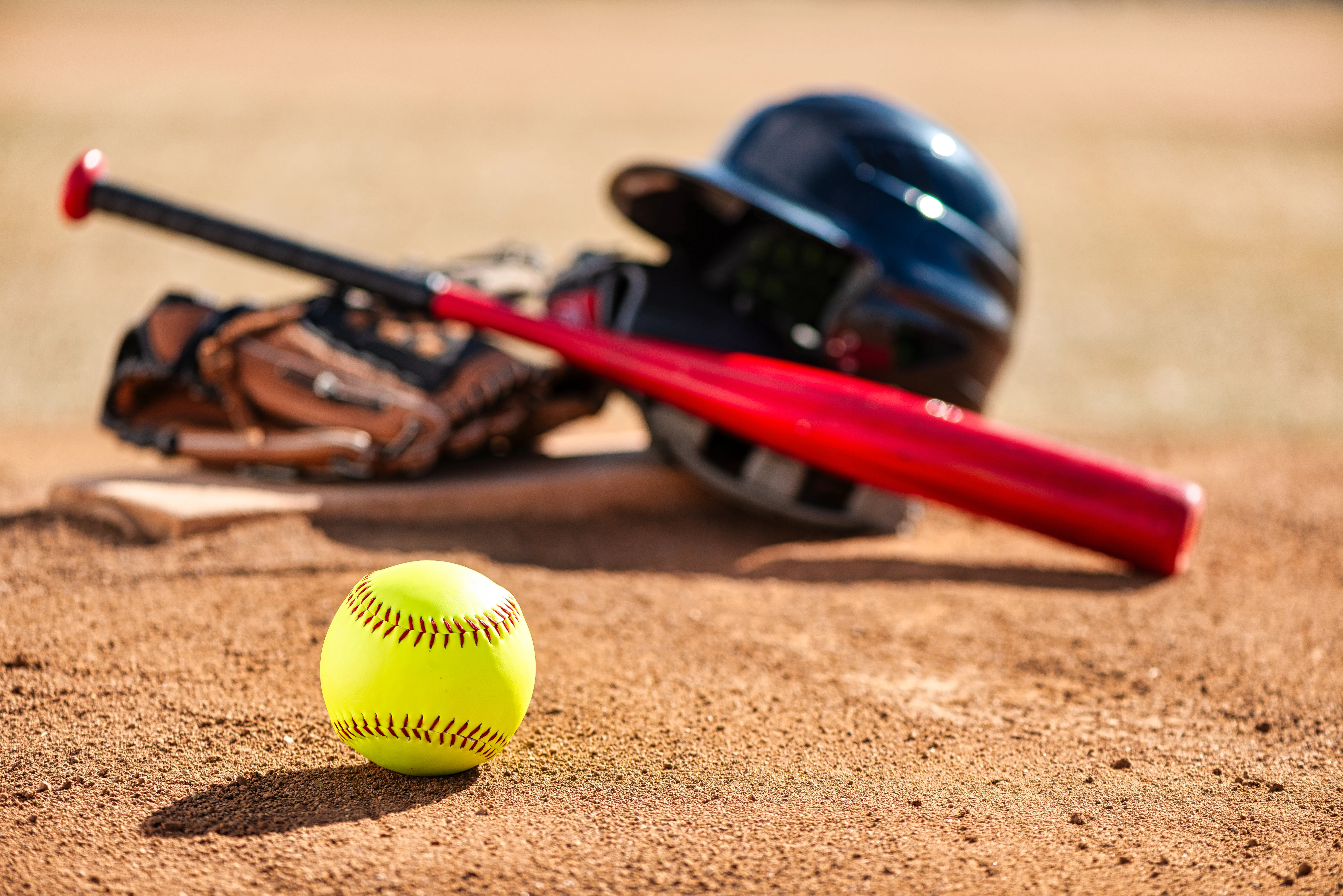 A softball sit in the foreground, with a helmet, bat, and glove sitting on a base slightly blurred in the background.