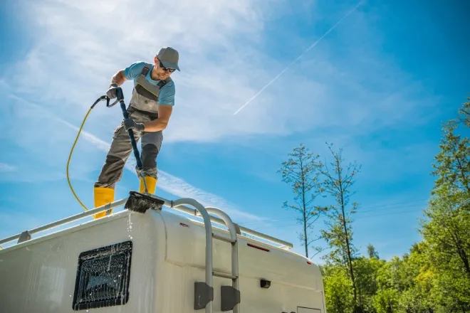A man in overalls and yellow rubber boots stands on top of an RV roof and cleans the vehicle using a powered brush.