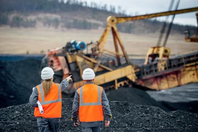 Two mining workers supervise a mechanical crane in a field of coal mounds. One of them gestures toward the machine.