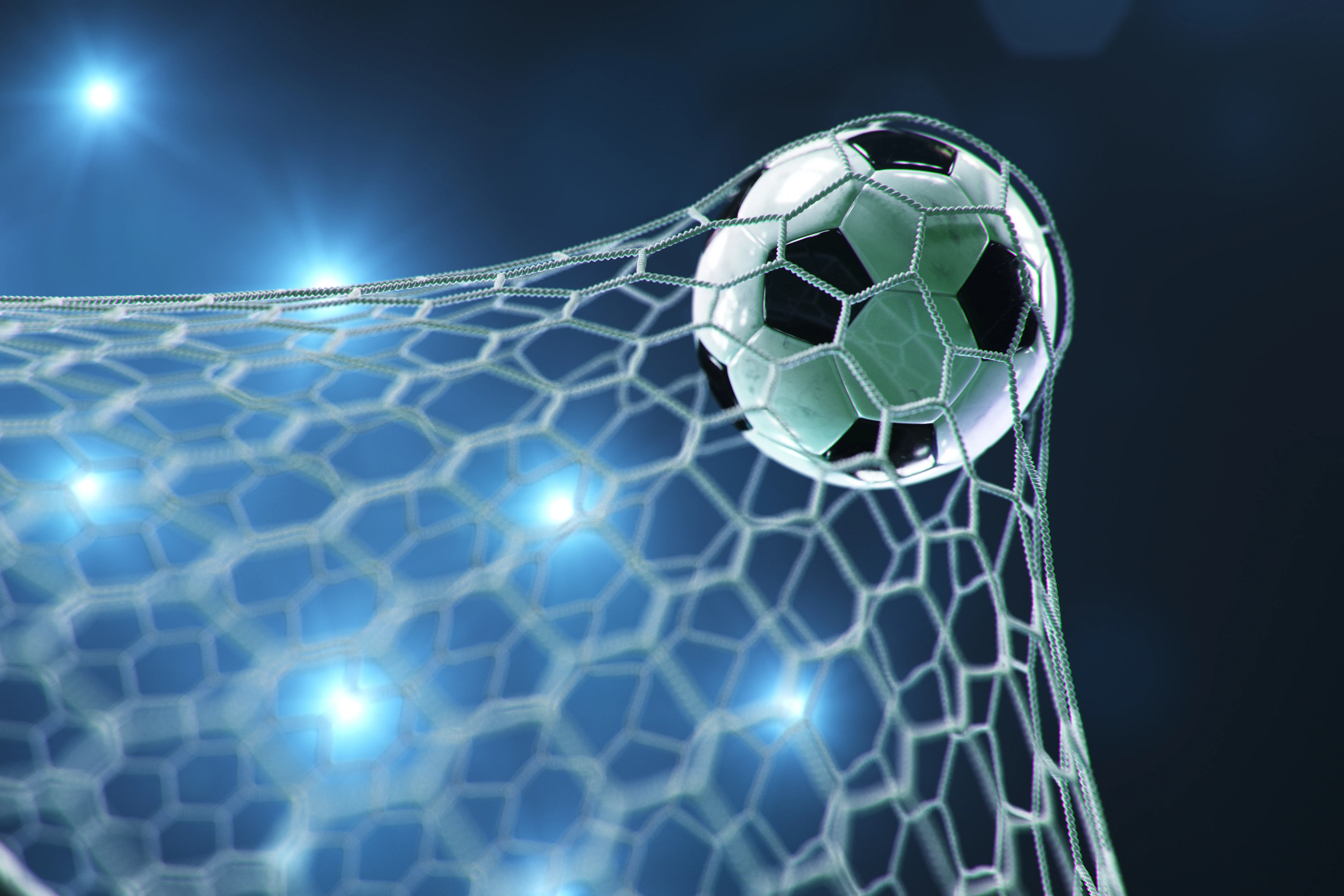 Soccer ball/football stretching the goal net with stadium lights in the background.