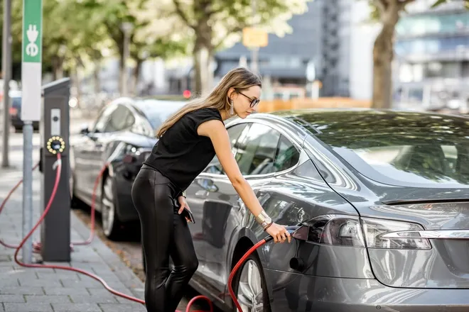 A young woman stands in front of her electric car on the side of a road. She plugs a connector into the car to charge it.