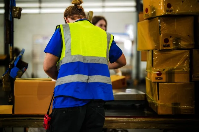 Two people working in a warehouse. They're packing boxes and stacking them together for shipment, wrapped tightly.