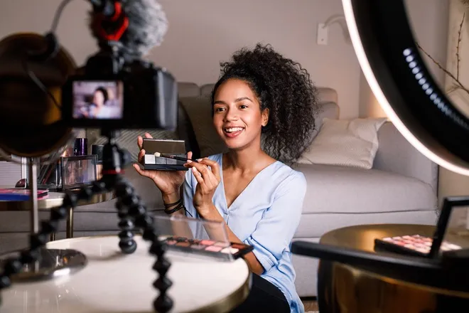 A young, Black influencer sits in front of a tripod and camera setup as she films a makeup eyeshadow palette.