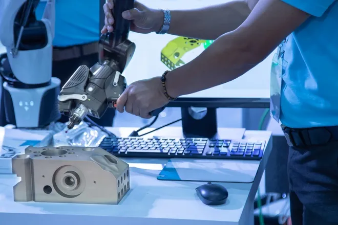 A technician stands in front of a portable 3D measuring arm. A part with grooves and holes rests on the table below the tool.