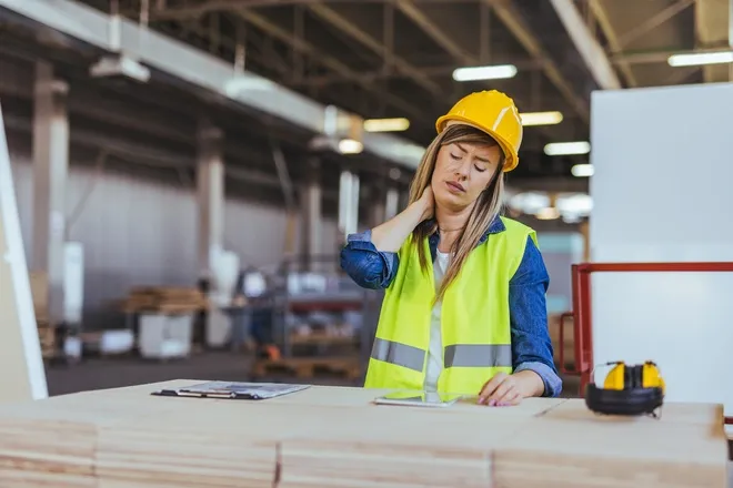 A female engineer sits at a work bench while she holds her neck in pain. She wears a hard hat and a safety vest.