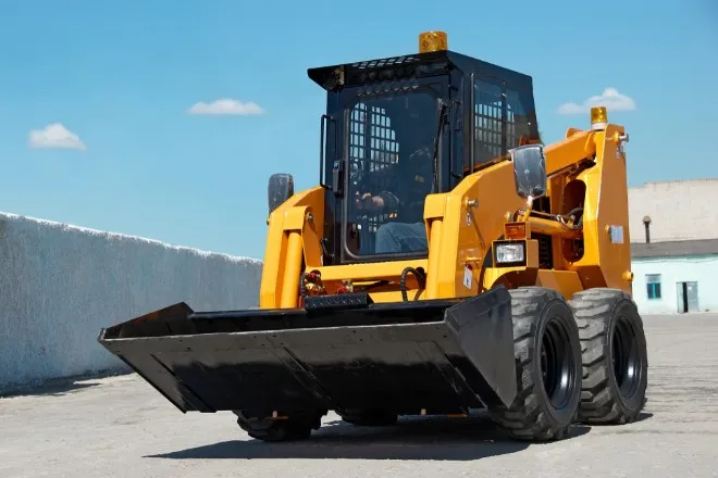 A yellow skid steer loader with a large black front bucket parked on a paved lot under a clear blue sky.