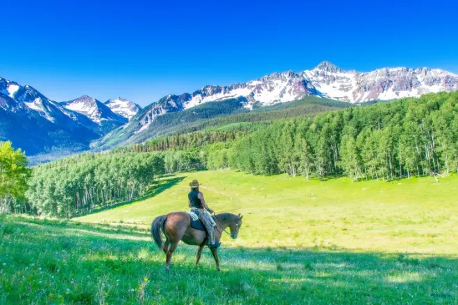 Cowboy riding a horse through a Colorado mountain landscape with grassy hills and alpine scenery in view.