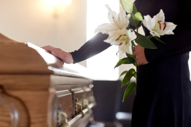 A woman stands in a black dress at a funeral. She holds white lilies in one hand and puts her other hand on the coffin.