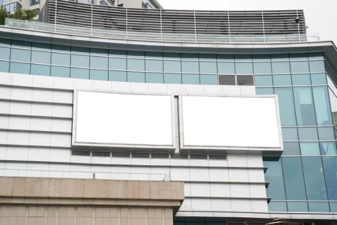 A low-angle view of two blank billboards hanging on the exterior of a large glass building on an overcast day.