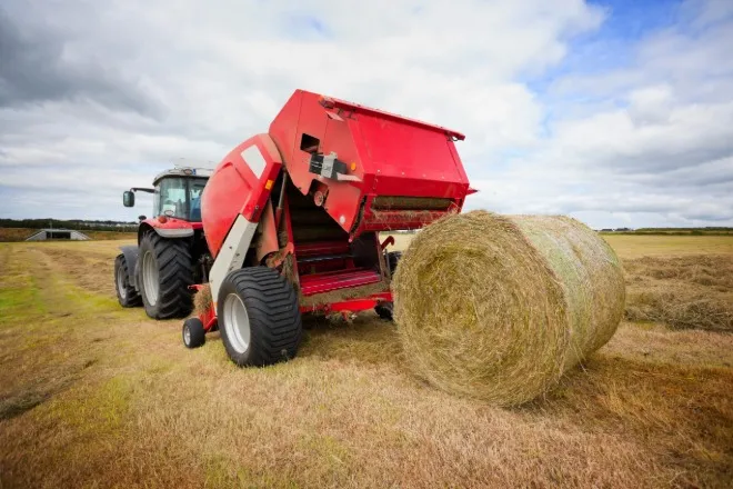  A rear view of a red farming tractor towing a red round hay baler as it drops a round hay bale in a field.