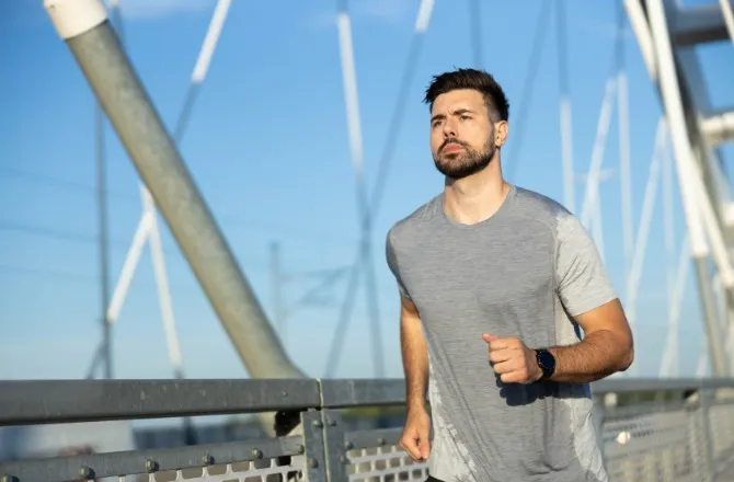 A man in a sweat stained shirt is jogging in an outdoor environment. Behind him is a suspension system for a bridge.