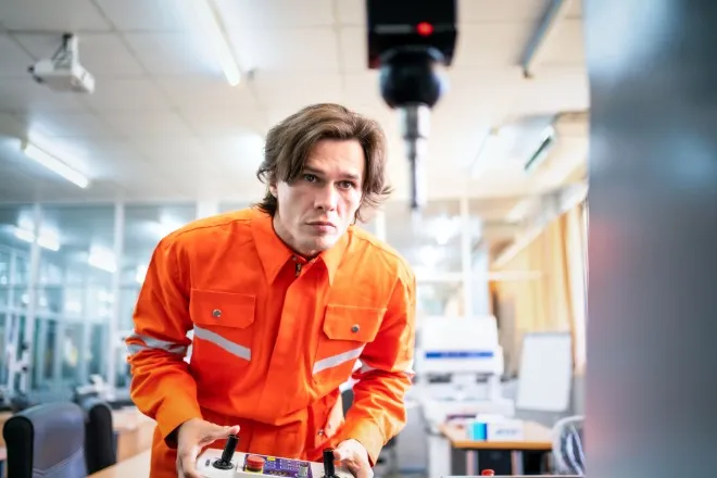 A CMM measurement probe is attached to a machine and blurred. A man wearing an orange work suit holds a controller.