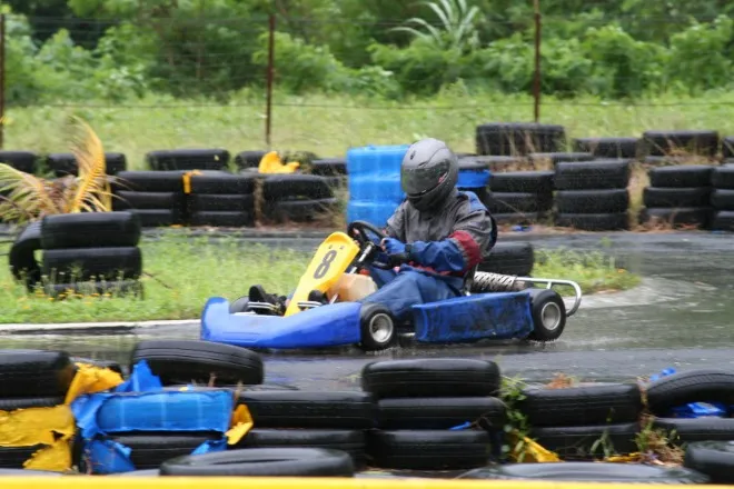 A driver navigates a tight corner of a go-kart track in the rain, with tires forming a safety barrier around them.