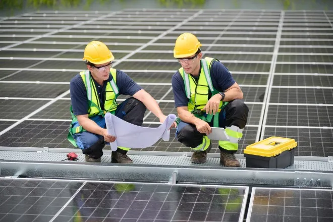 Two people standing near solar panels, holding and looking at blueprints while examining the installation site.