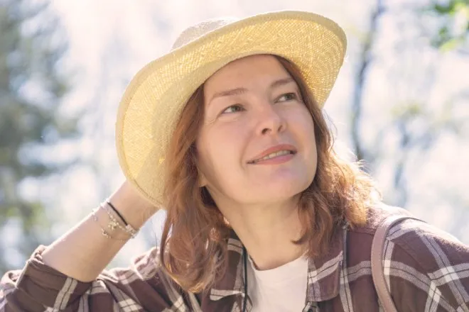 A woman wearing a straw hat and plaid shirt looking upward outdoors with trees and bright sunlight behind her.