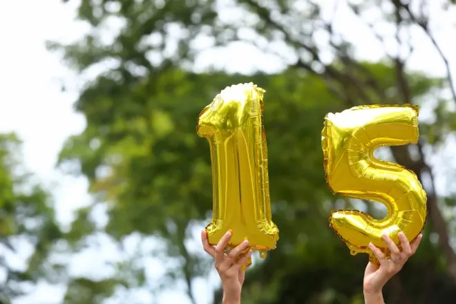 Two hands hold shiny gold number balloons forming 15 outdoors, with leafy green trees blurred in the background.