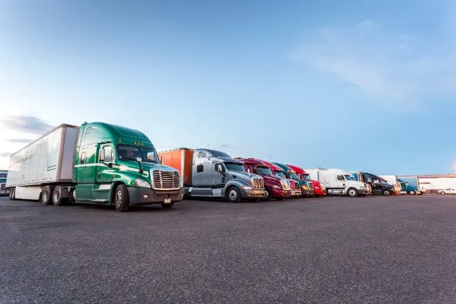 A row of colorful semi-trucks and trailers parked side by side in a large paved lot beneath a clear blue sky.