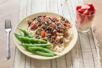 Plate on a placemat of picadillo on a bed of rice with green beans. A fork and glass of sliced strawberries are next to the plate.