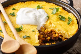 Skillet of stovetop tamale pie topped with sour cream with two wooden serving spoons and a slice removed.