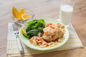 Table with a placemat under a plate of chicken and rice with spinach leaves and a lemon wedge next to a fork, glass of milk, and bowl of lemon or orange wedges.