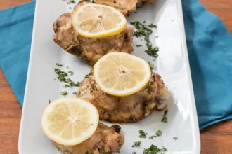 Serving platter sitting on a cloth on a wooden table, with three pieces of citrus chicken topped with slices of citrus on top.