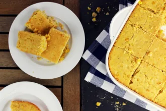 Plate with three squares of corn bread next to a baking dish of corn bread on a cloth.