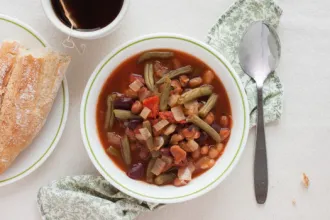 Bowl of Italian Bean Soup on a cloth. Near by are a spoon, cup of hot tea, and plate with bread.