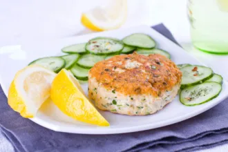 Plate containing a lemon pepper salmon patty, lemon wedges, and slices of cucumber.