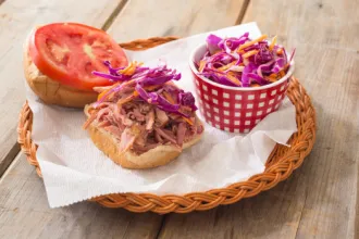 Pulled Pork Sandwich with Red Cabbage and Carrot Slaw - USDA Wicker basket lined with a paper towel. A container of red cabbage and carrot slaw sits to one side, accompanied by a bread bun with pulled pork on one half and a tomato slice on the other half.