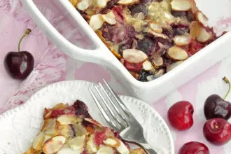 Overhead view showing part of a baking dish of cherry puff pancake and part of a plate with a fork and a serving of the pancake.